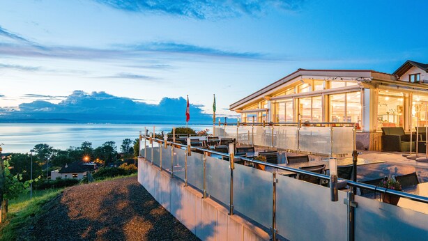 Un hôtel situé au bord de l’eau, doté d’une façade en verre, de places assises en plein air et offrant une vue sur l’eau.