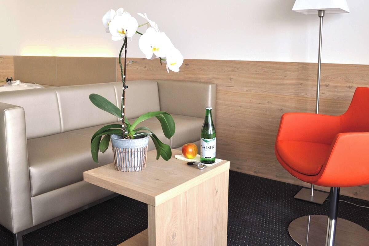Habitació d'hotel amb un sofà, una taula de fusta petita amb una orquídia en test, una ampolla de VAISE, una poma i una cadira taronja moderna.