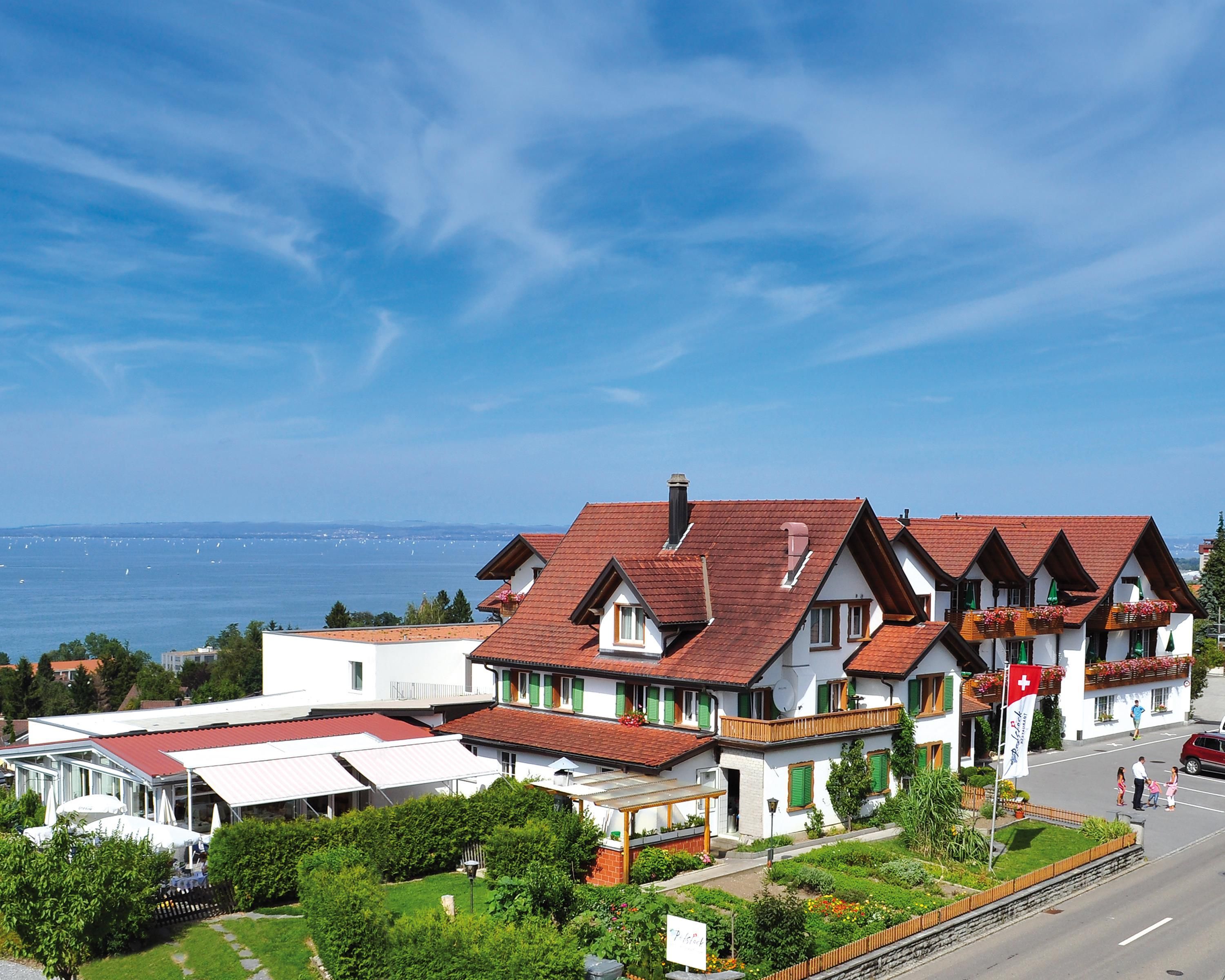 Il s’agit d’un bâtiment de style chalet traditionnel, avec un toit recouvert de tuiles rouges, des murs blancs et des volets verts, situé près d’une étendue d’eau.
