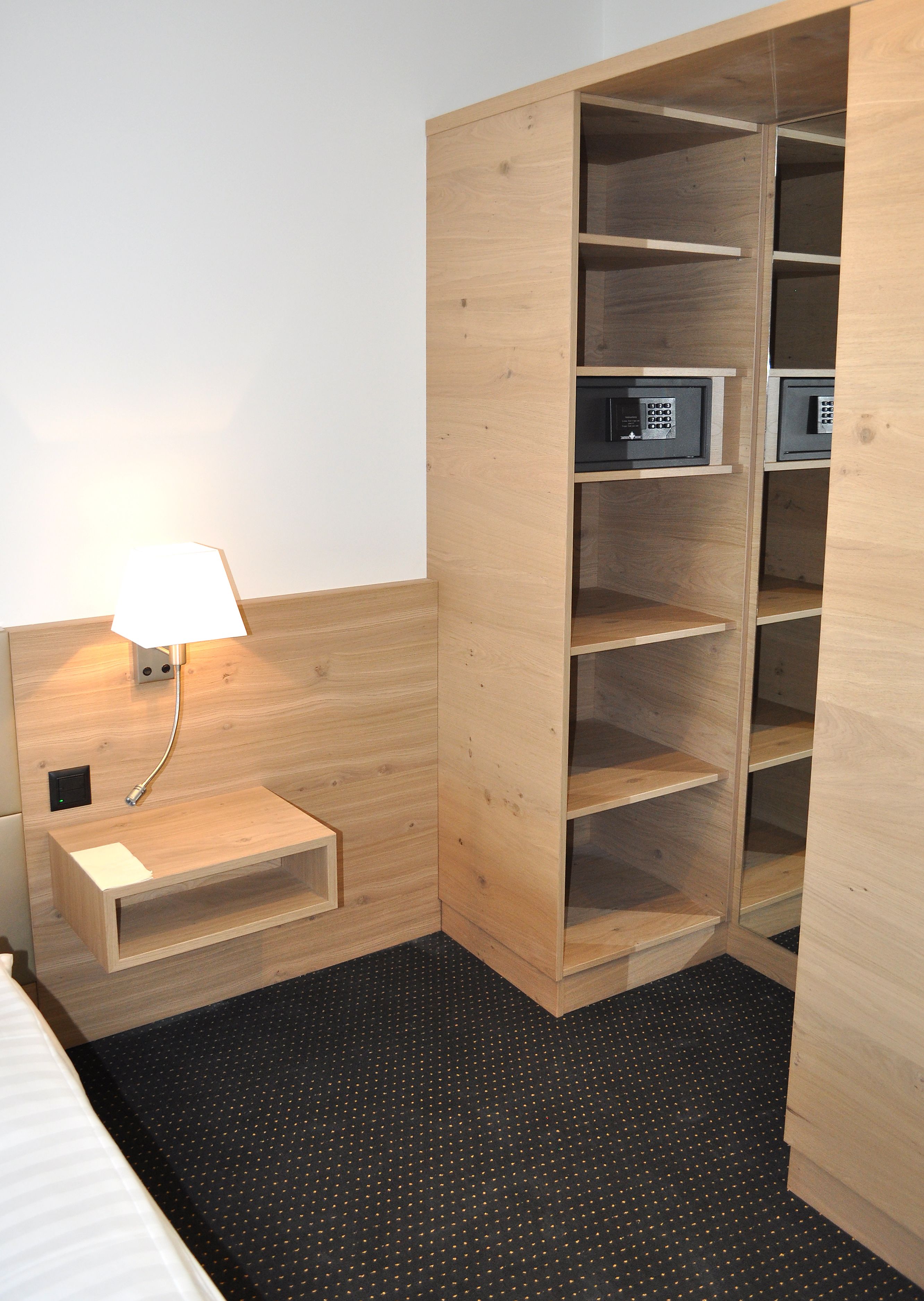 A hotel room with a wooden wardrobe, a bedside table with a lamp, and a black carpet with a dotted pattern.