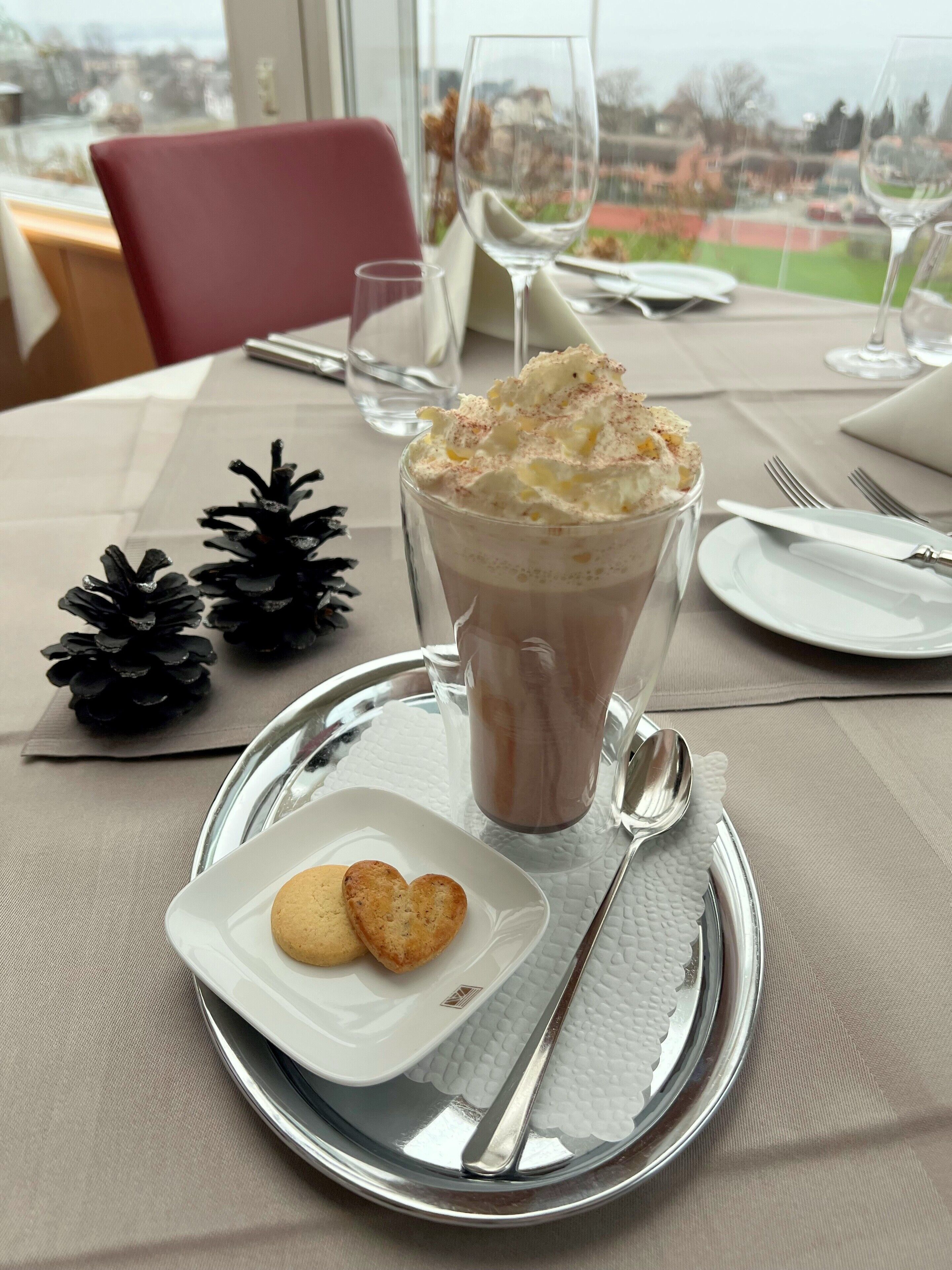 Una mesa de comedor con un postre: un vaso de batido de chocolate cubierto de crema batida y una galleta en un plato pequeño.