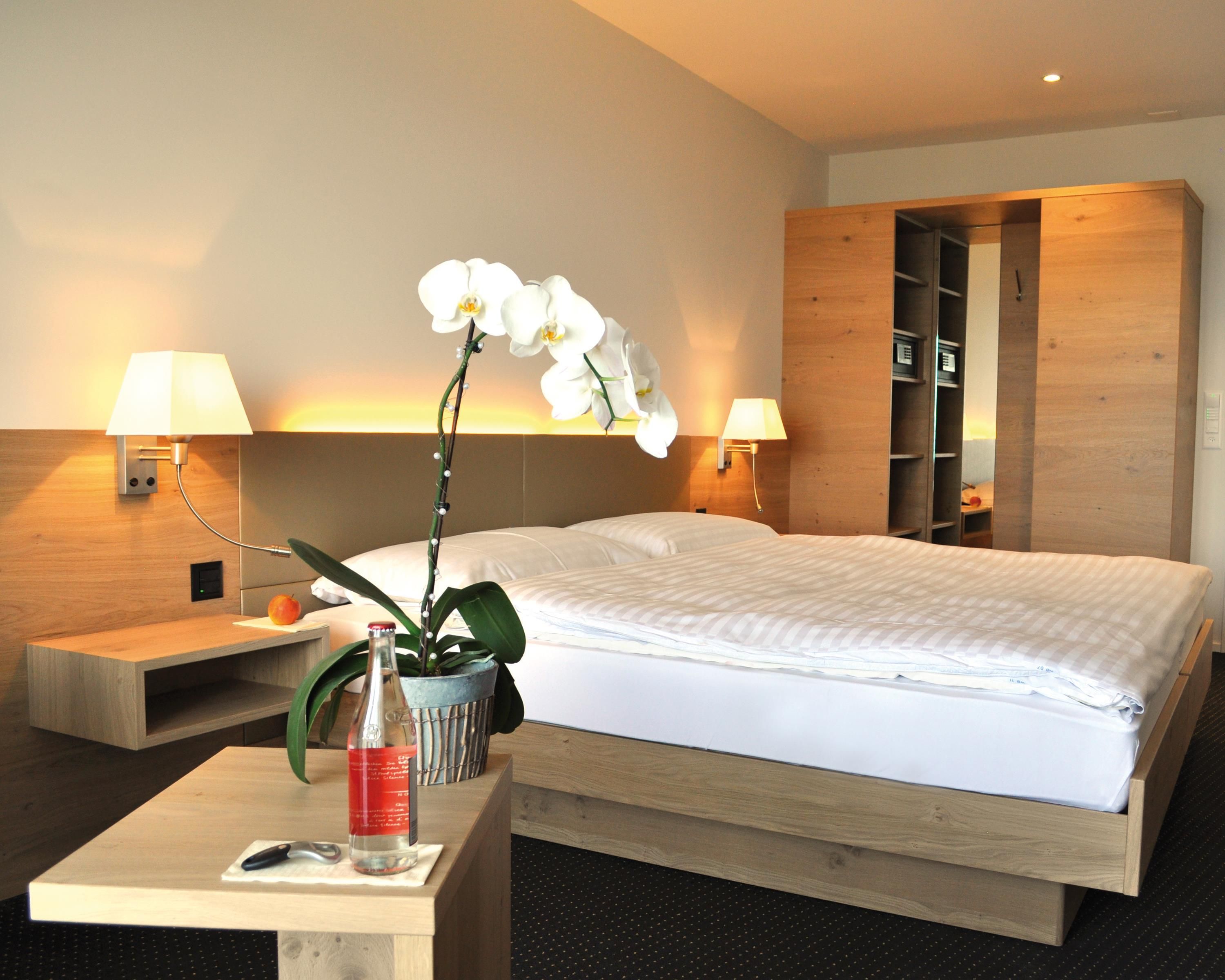 A hotel room with a bed, two bedside lamps, a nightstand with a bottle and a fruit, and a small table with a bottle and a knife.