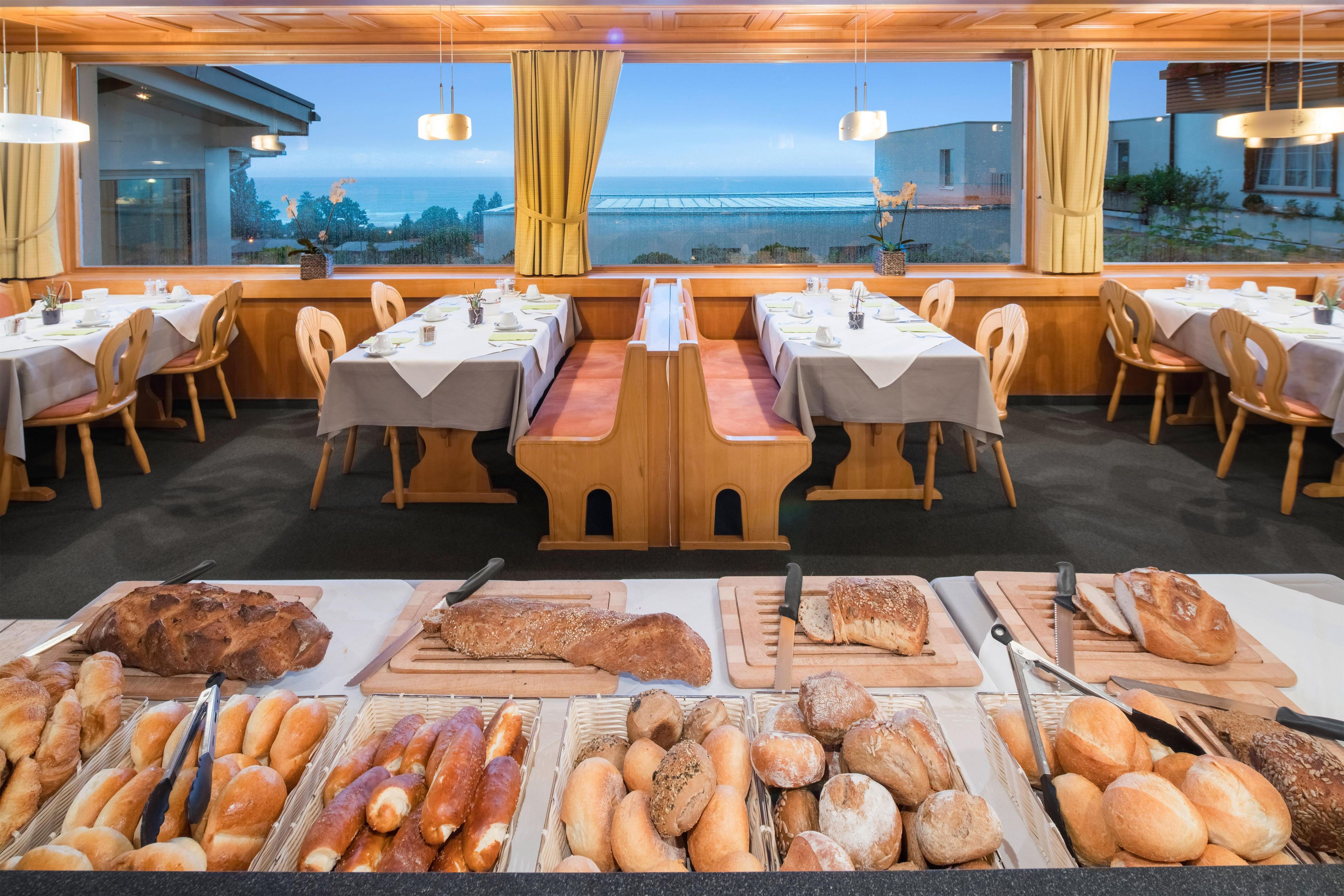A restaurant with tables set for dining, a variety of bread displayed in the foreground, and a view of the sea through large windows.