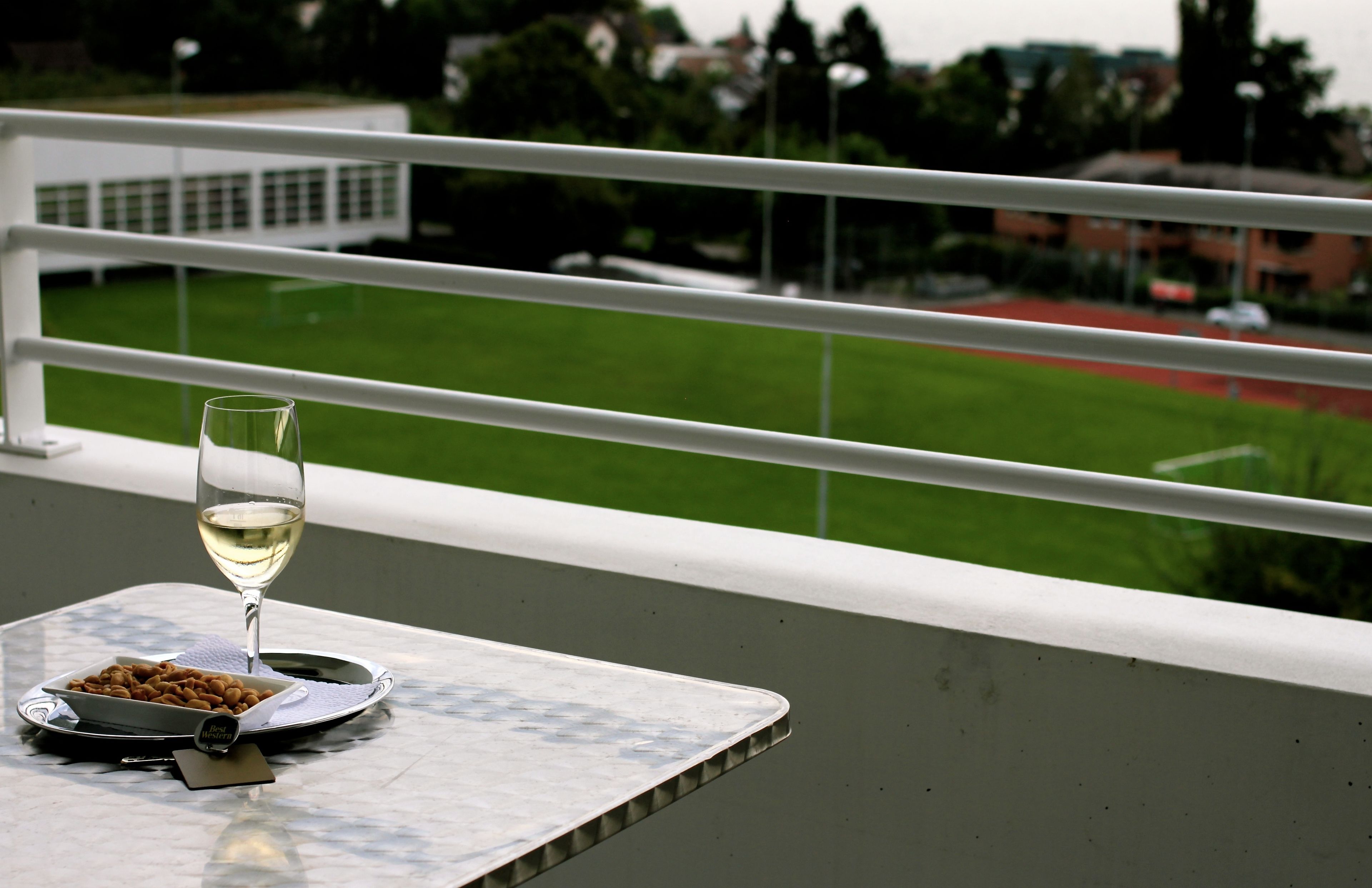 Een tafel met een glas witte wijn en een bord amandelen, met uitzicht op een groen veld en gebouwen.