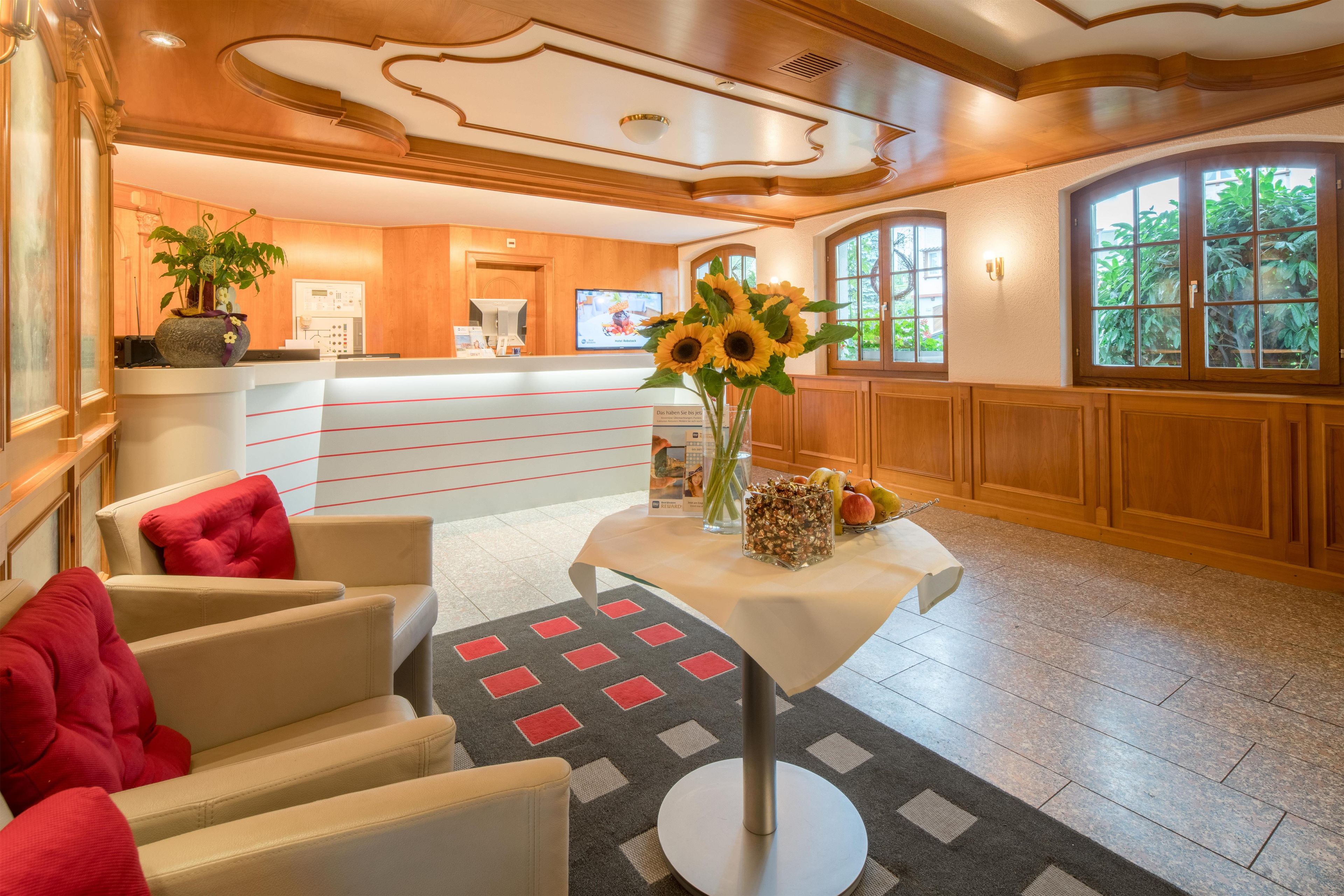 A hotel lobby with a reception desk, a seating area with red cushions, a vase with sunflowers, and a small table with a decorative item.