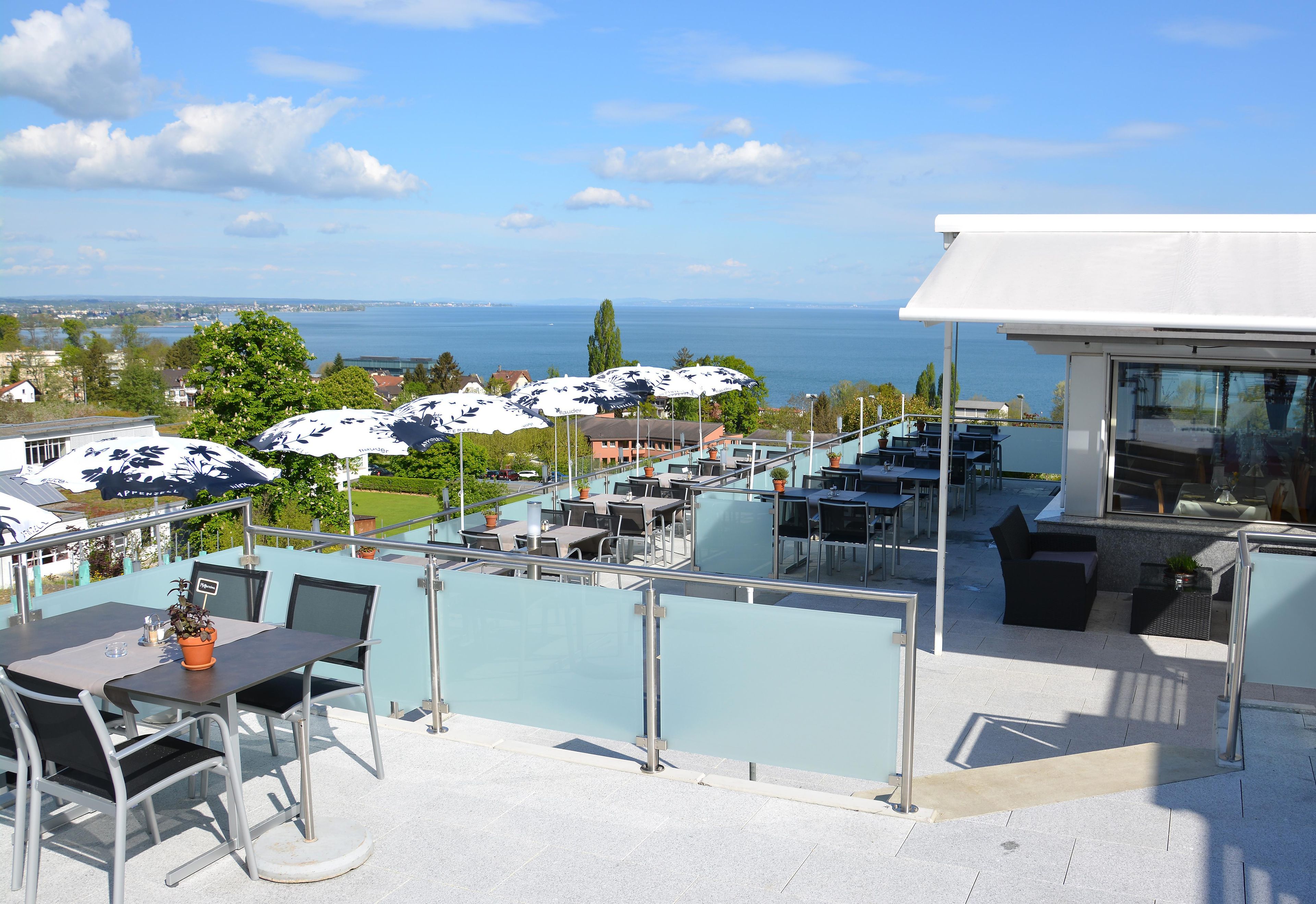 Um terraço ao ar livre com mesas, cadeiras, guarda-sóis e vista para o mar.