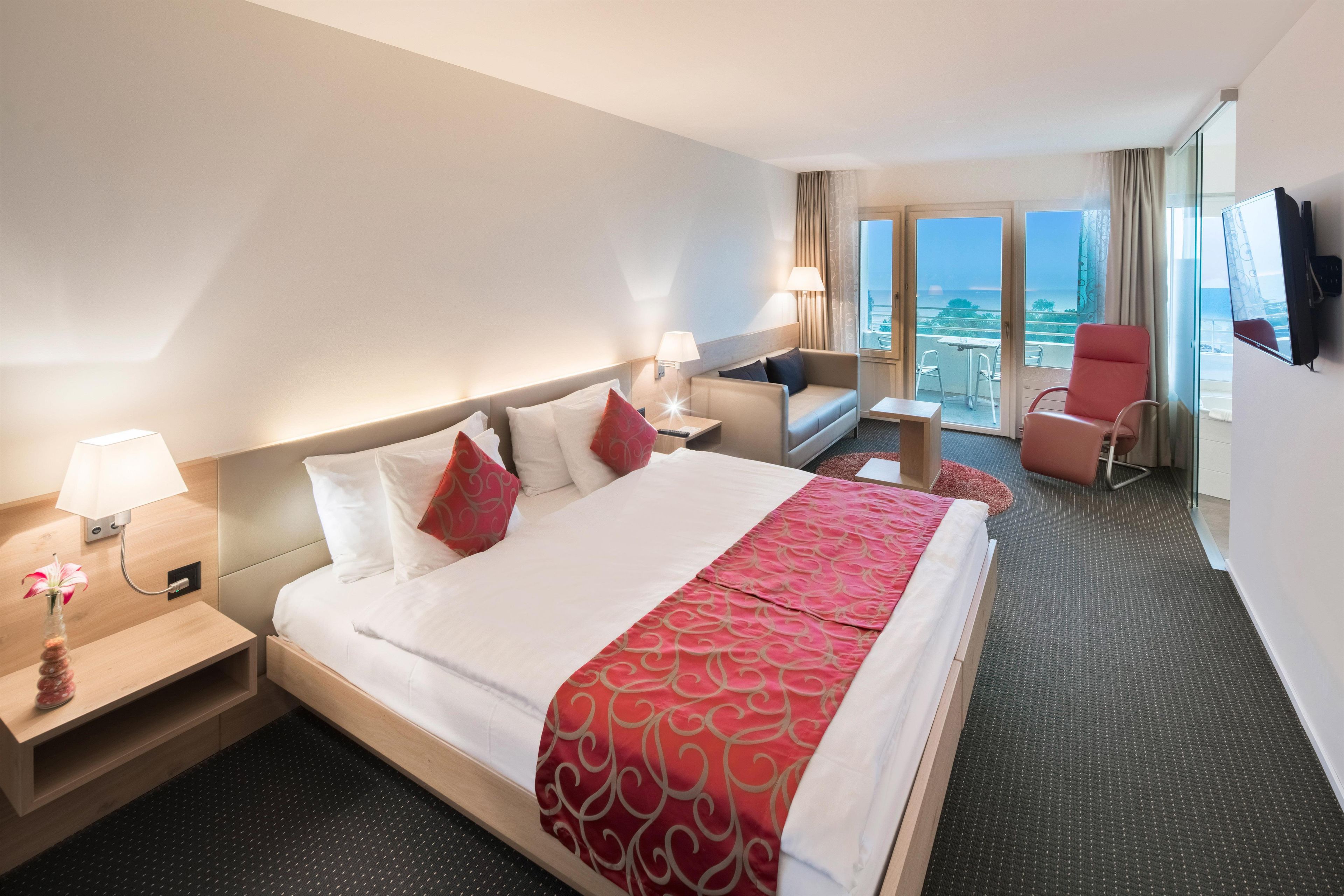 A hotel room with a large bed, a desk, a chair, a TV, and a balcony with a view.