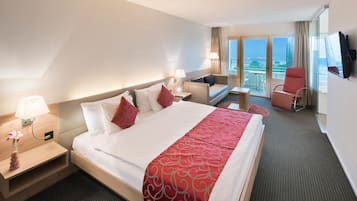 A hotel room with a large bed, a desk, a chair, a TV, and a balcony with a view.