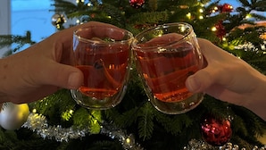Un árbol de Navidad decorado con adornos y luces, y dos manos sosteniendo vasos con líquido rojo.