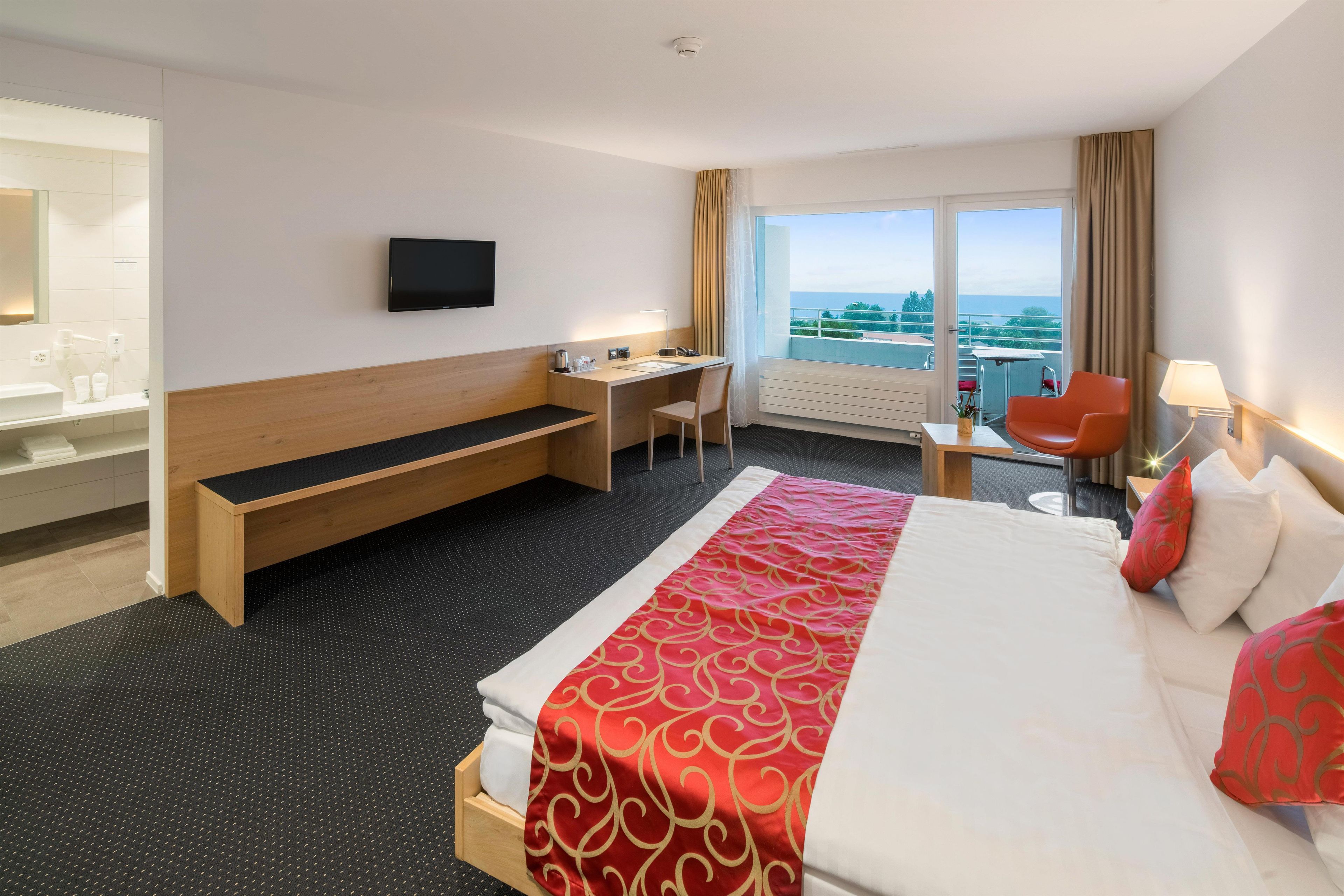 A hotel room with a bed, a desk, a chair, a TV, and a view of the sea.