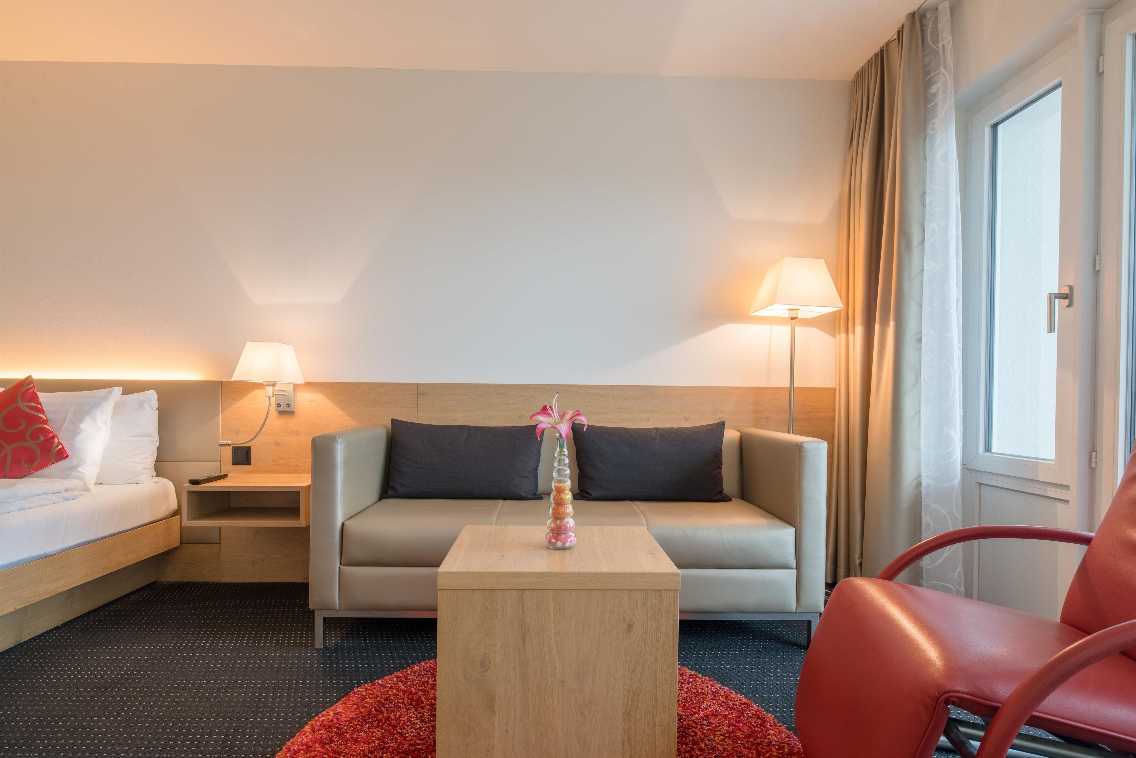Una habitación de hotel con una cama, un sofá, una mesita y una silla. Hay dos lámparas de noche y una ventana con cortinas.