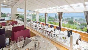 Un restaurant où les tables sont dressées pour le service, avec de grandes fenêtres offrant une vue sur la mer et les bâtiments, et un décor intérieur moderne.