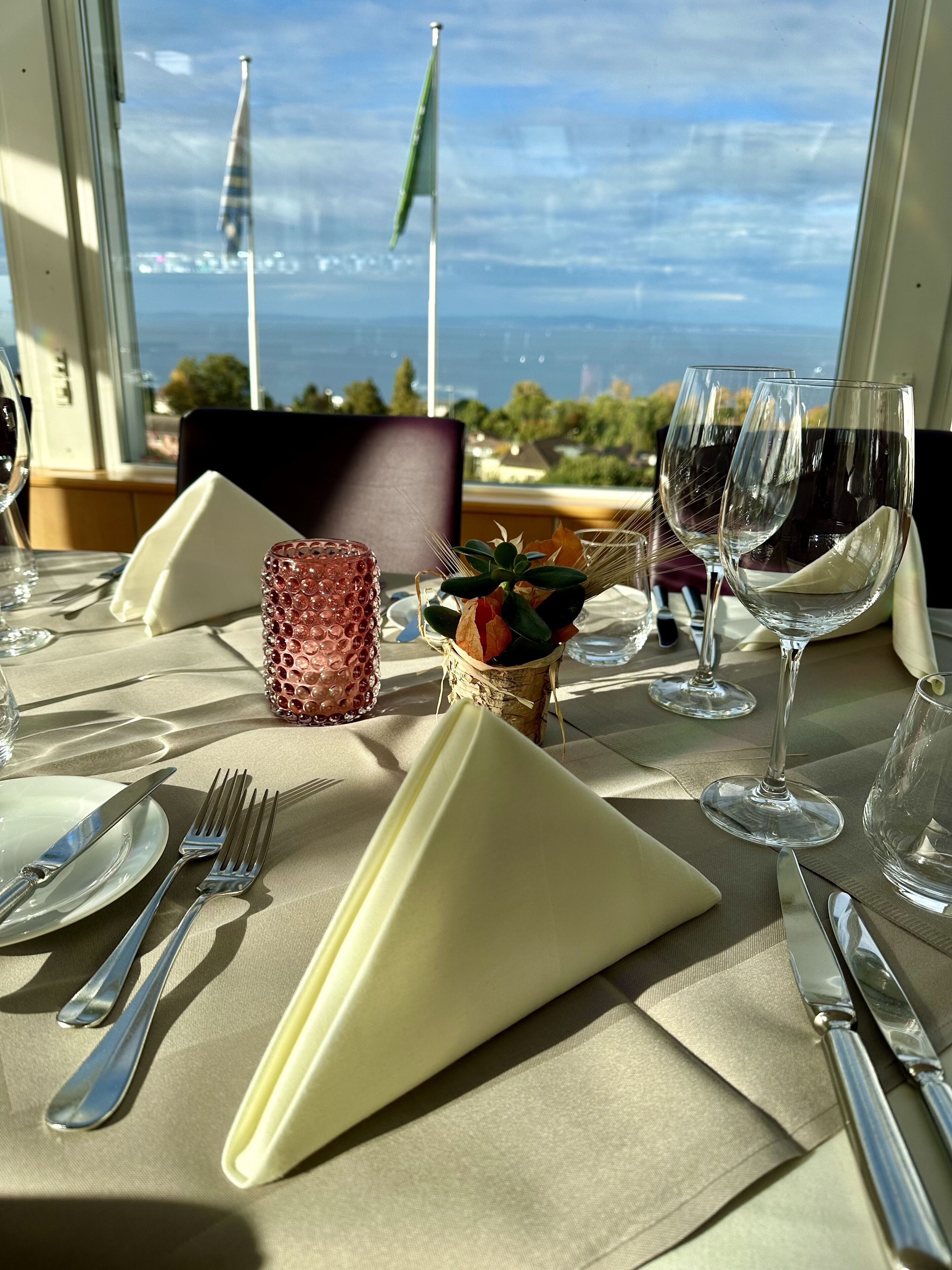 A table set for dining with a view of the sea and flags, featuring a vase with dried leaves and a textured glass candle holder.