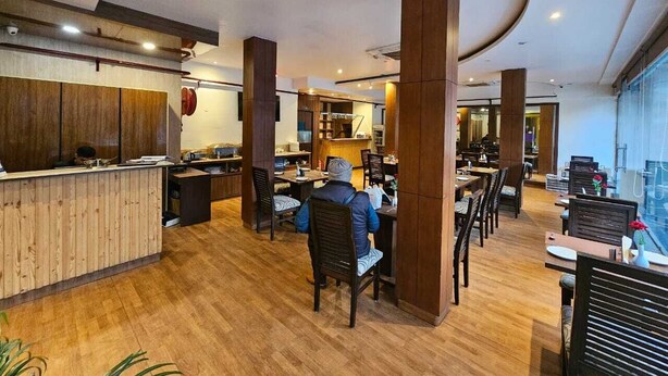 A modern restaurant with wooden flooring and high ceilings, featuring a central dining area with tables and chairs, and a reception desk. - Hotel Saptagiri (New Delhi)