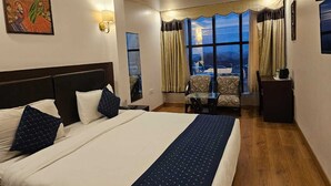 A hotel room with a large bed, a desk, a chair, and a view of the city through large windows. - Hotel Saptagiri (New Delhi)