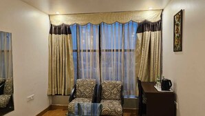 A living room with two patterned armchairs, a glass coffee table, a dark wooden side table, and a window with sheer curtains. - Hotel Saptagiri (New Delhi)