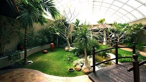 A lush indoor garden with a variety of plants, a small pond, and a wooden deck. - Celestia Suites at Leonia (Shamirpet)
