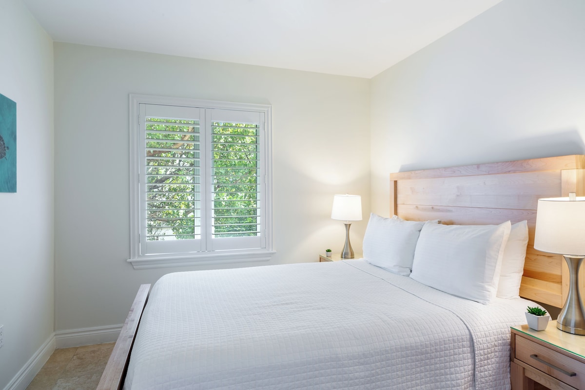 A neatly made bed with white linens, a bedside table with a lamp, and a window with blinds.