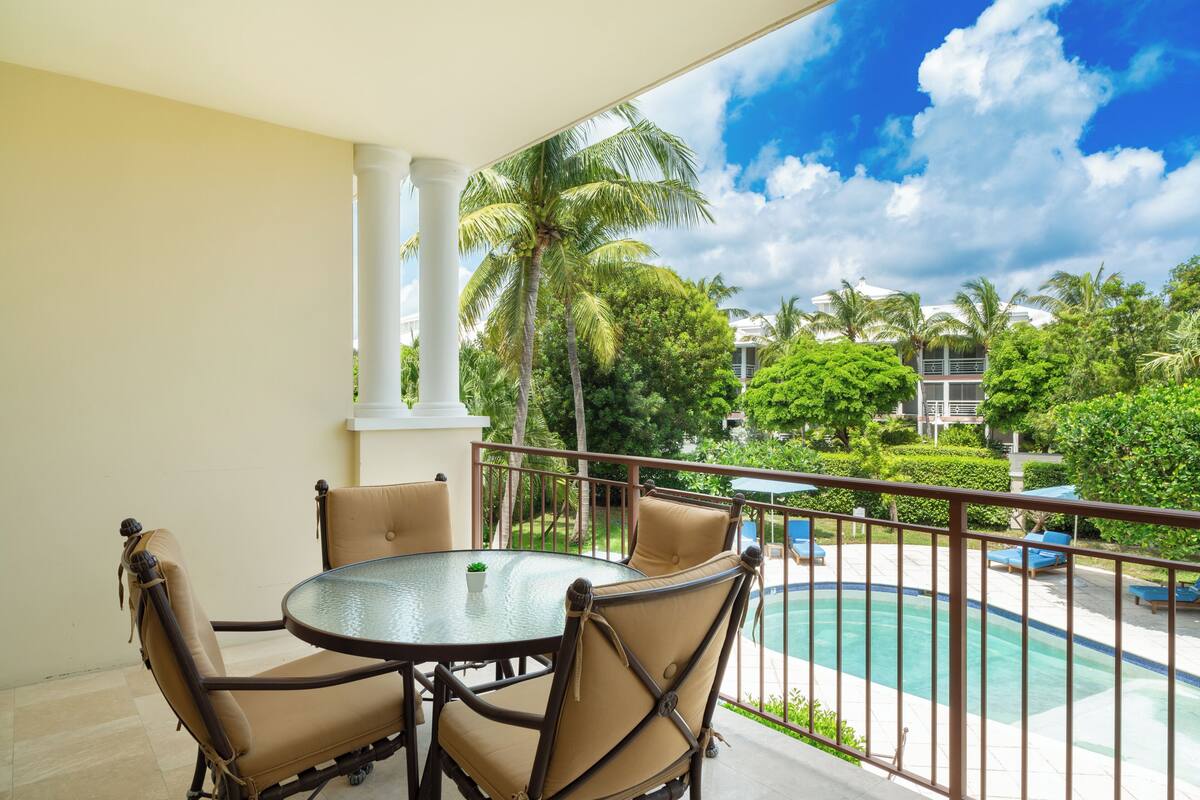 A balcony with a glass table and four chairs, overlooking a swimming pool and lush greenery.