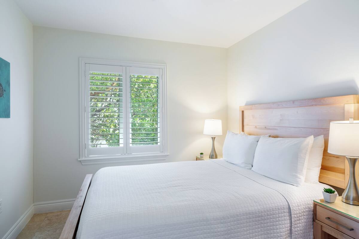 A neatly made bed with white linens, a bedside table with a lamp, and a window with blinds.