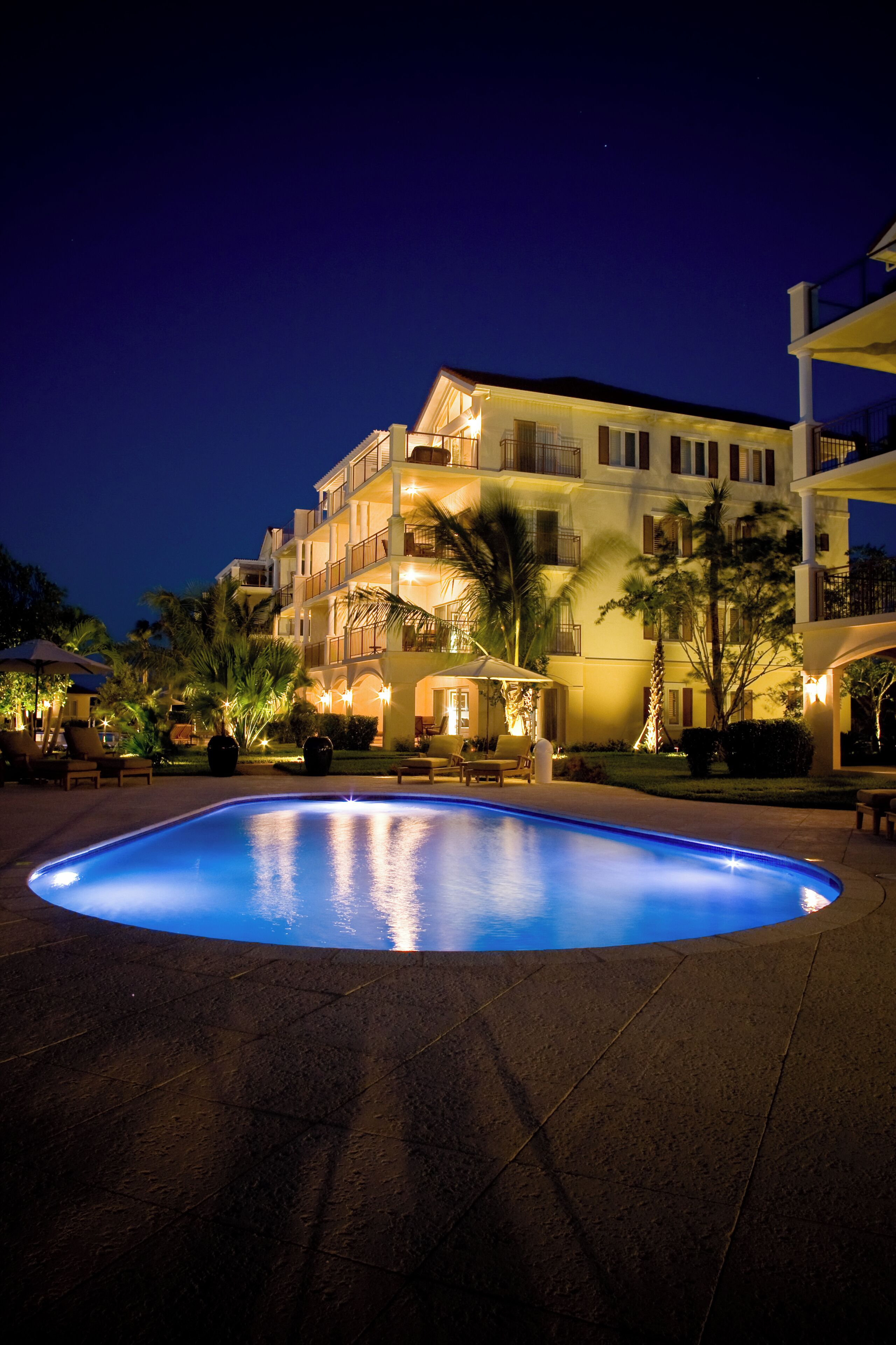 Un área de piscina iluminada durante la noche, con un edificio al fondo.
