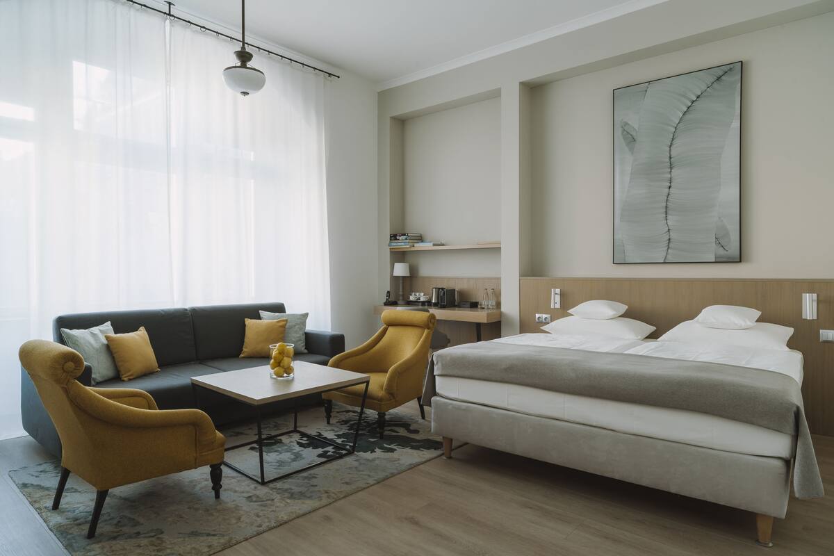 Una habitación de hotel moderna con sofá, dos sillones, una mesita de café, una cama y un cuadro abstracto grande en la pared.