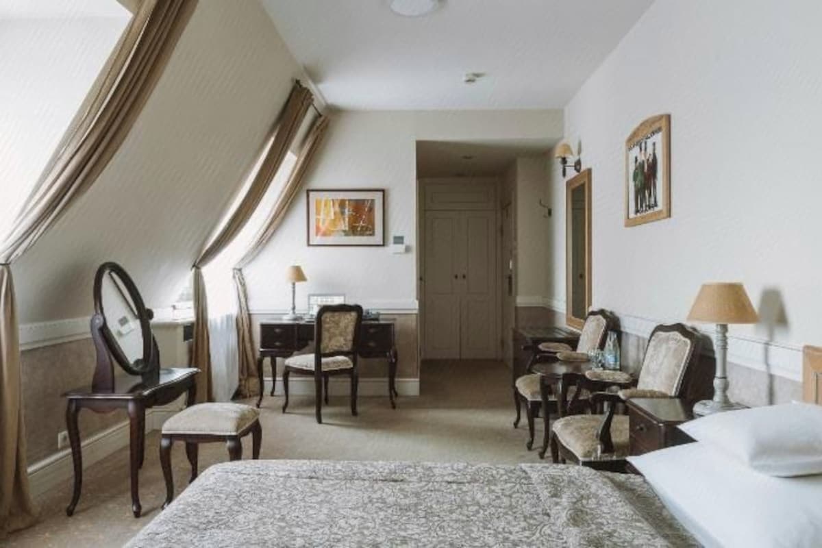 A hotel room with a large bed, a desk, a chair, and a small table. There is a door leading to another room.