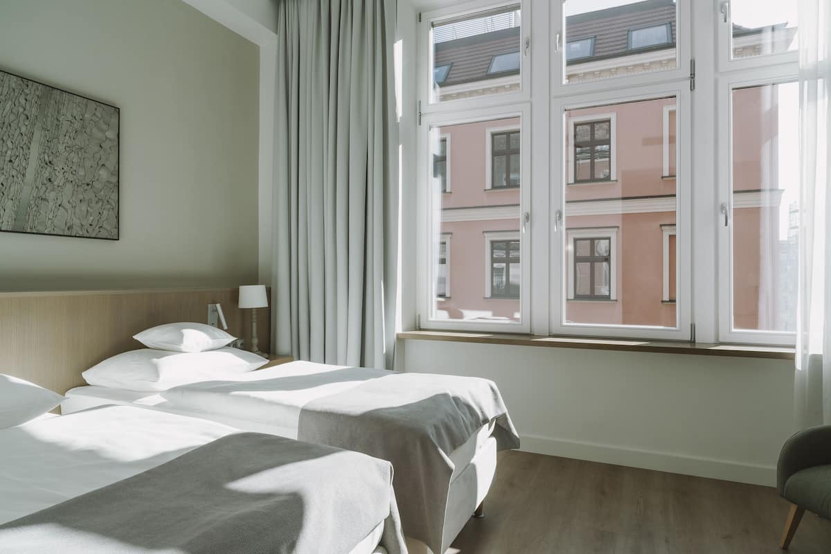 Habitación de hotel con dos camas, una ventana con vista a edificios, un cabecero y una obra de arte colgada en la pared.