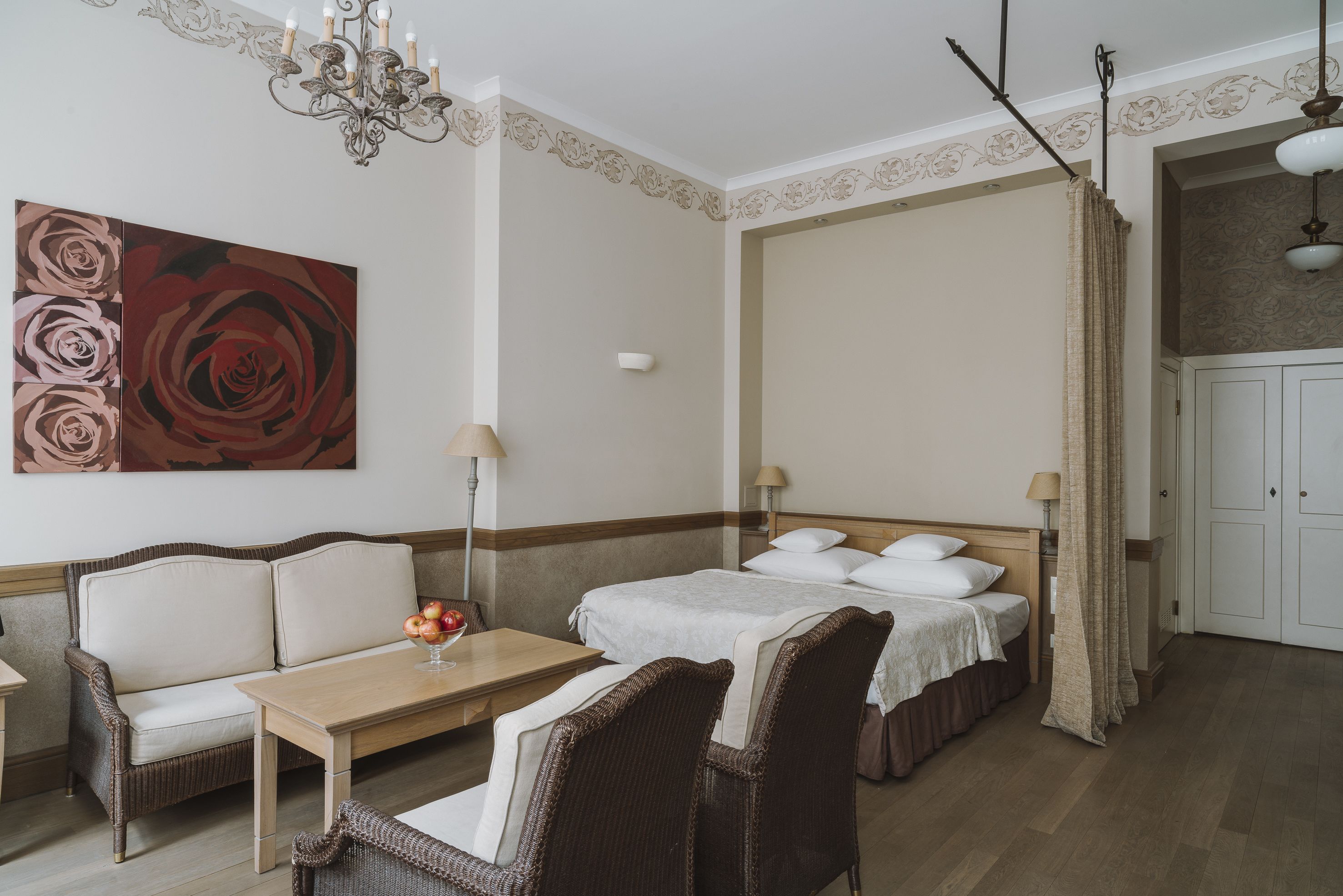 Een hotelkamer met een bank, twee enkele bedden, een tafel met een schaal fruit, een kroonluchter en een groot bloemenportret.