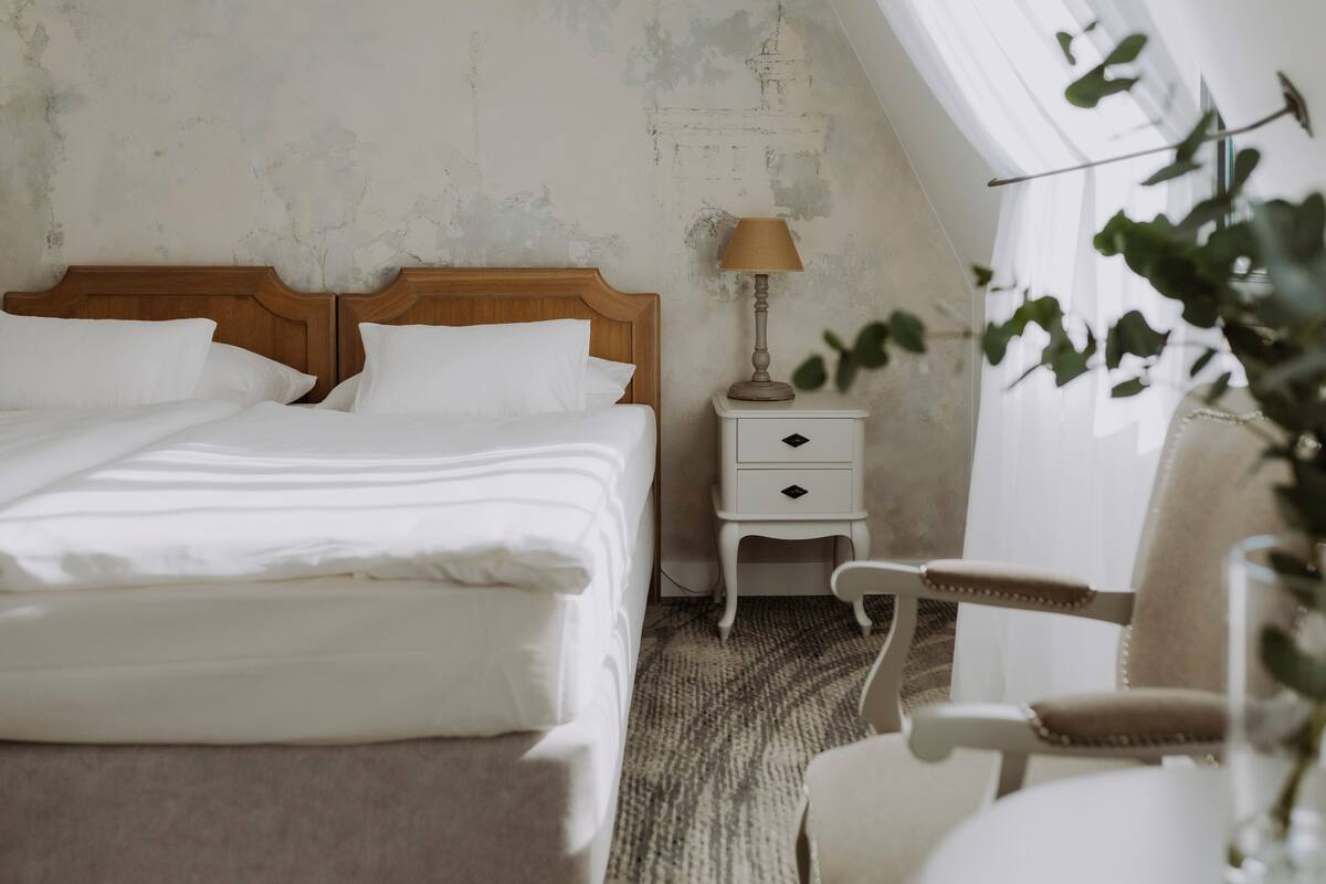 Un dormitorio con cabecera de madera, una mesita de noche blanca con lámpara, una silla y una planta.