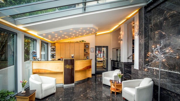 A modern reception area with a wooden reception desk, white leather chairs, and a glass ceiling. - Hotel Spitzberg (Passau)