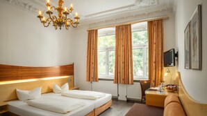 A hotel room with a wooden headboard, a large bed with white linens, a bedside table with a lamp, a TV mounted on the wall, and a window with curtains. - Hotel Spitzberg (Passau)