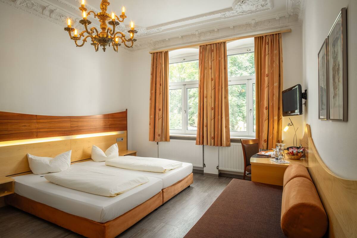 Une chambre d’hôtel avec une tête de lit en bois, un grand lit avec du linge de lit blanc, une table de chevet avec une lampe, un téléviseur fixé au mur et une fenêtre avec des rideaux.