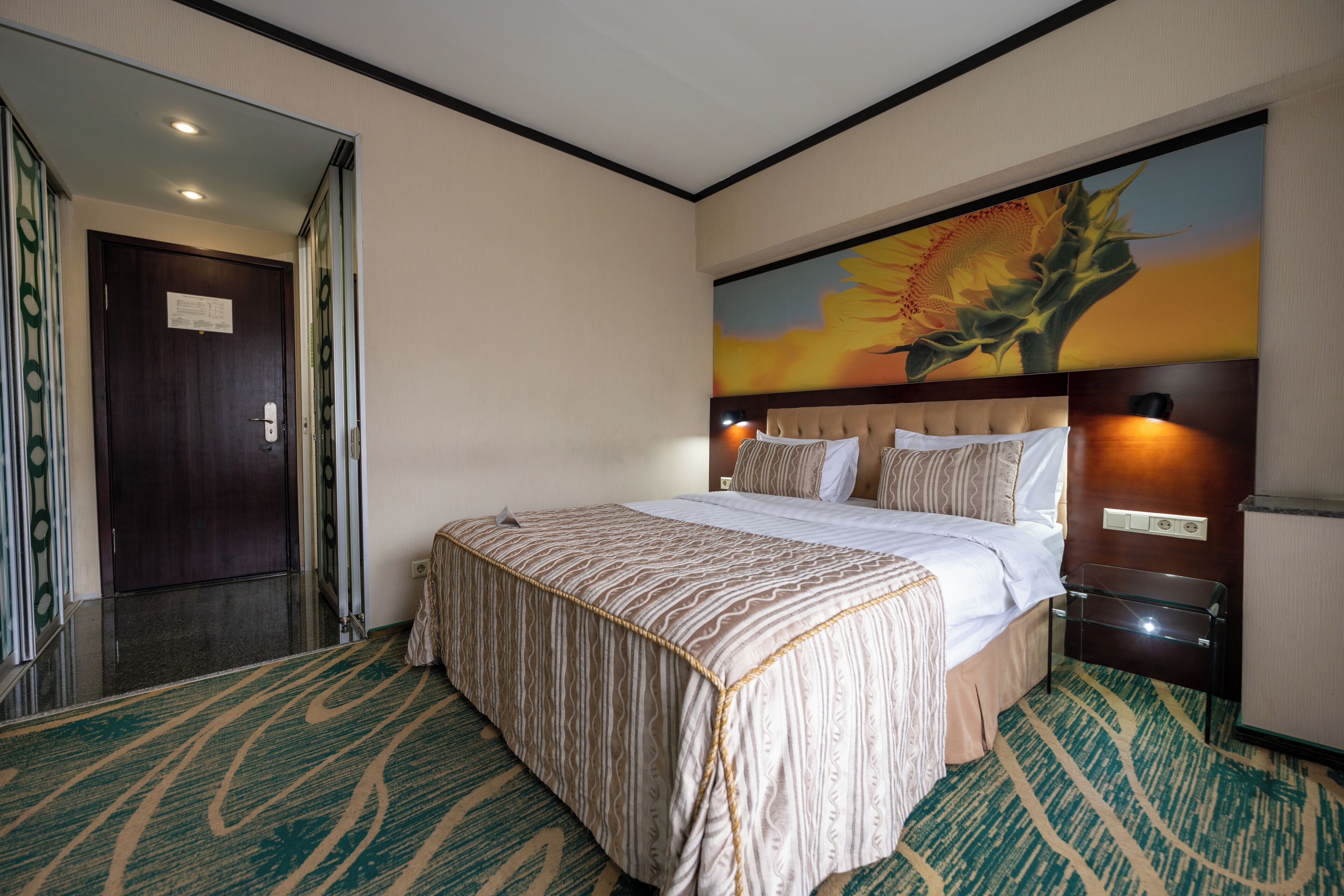 A hotel room with a large bed, a headboard with a floral mural, a glass side table, and a carpeted floor.