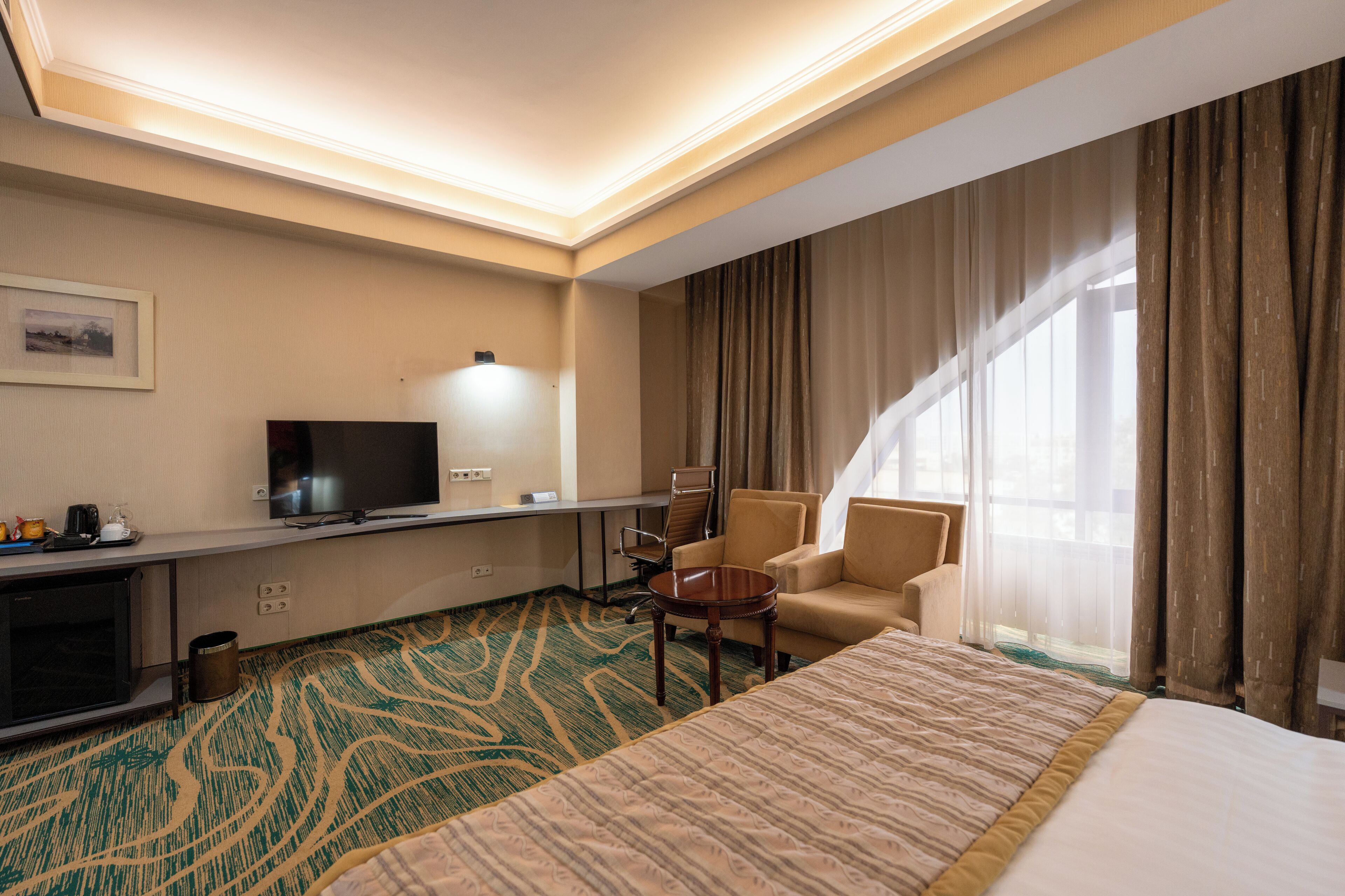 A hotel room with a large bed, a desk with a flat-screen TV, a chair, and a small table.