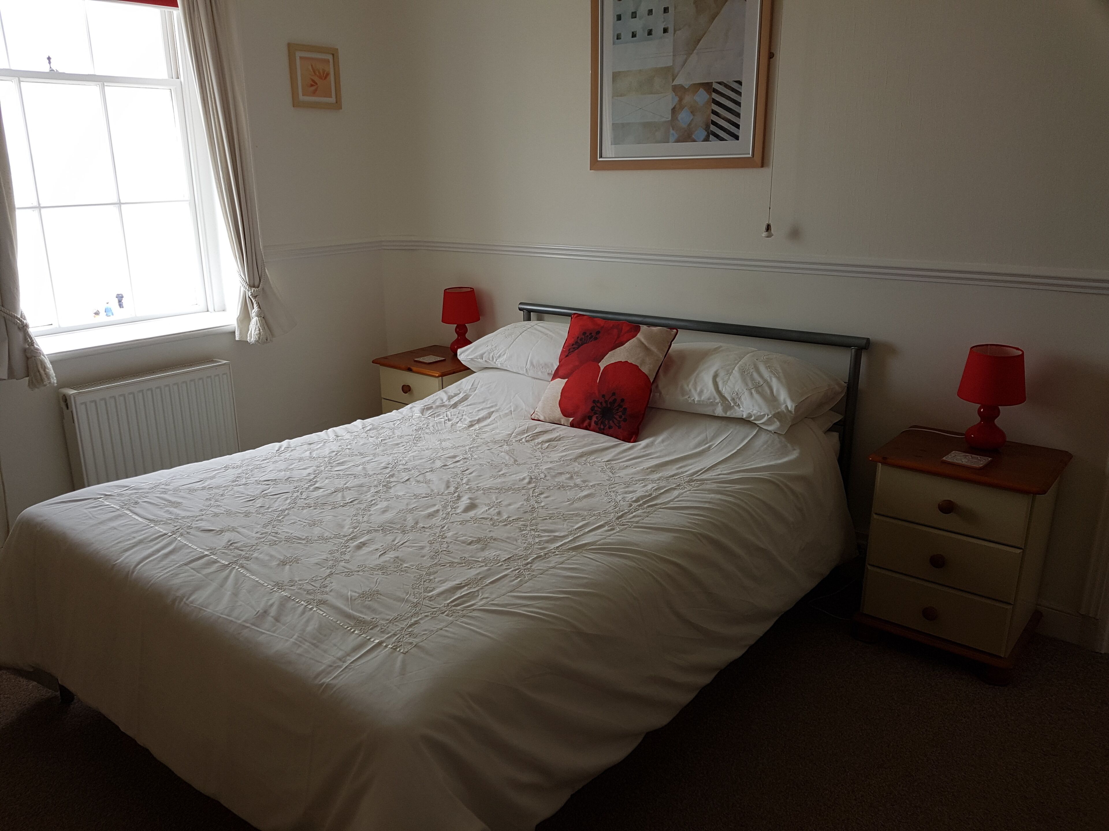 A neatly made bed with a white duvet, two bedside tables with red lamps, and a framed picture on the wall.