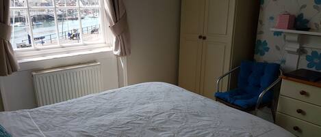 Et soveværelse med seng, stol, garderobe, radiator og et vindue med udsigt over havnen.