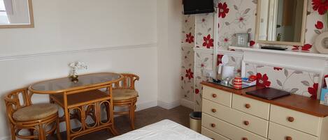 Une chambre d’hôtel comprenant un lit, une table en bois avec des chaises, une commode avec des tiroirs, un téléviseur fixé au mur et un papier peint à motifs floraux.