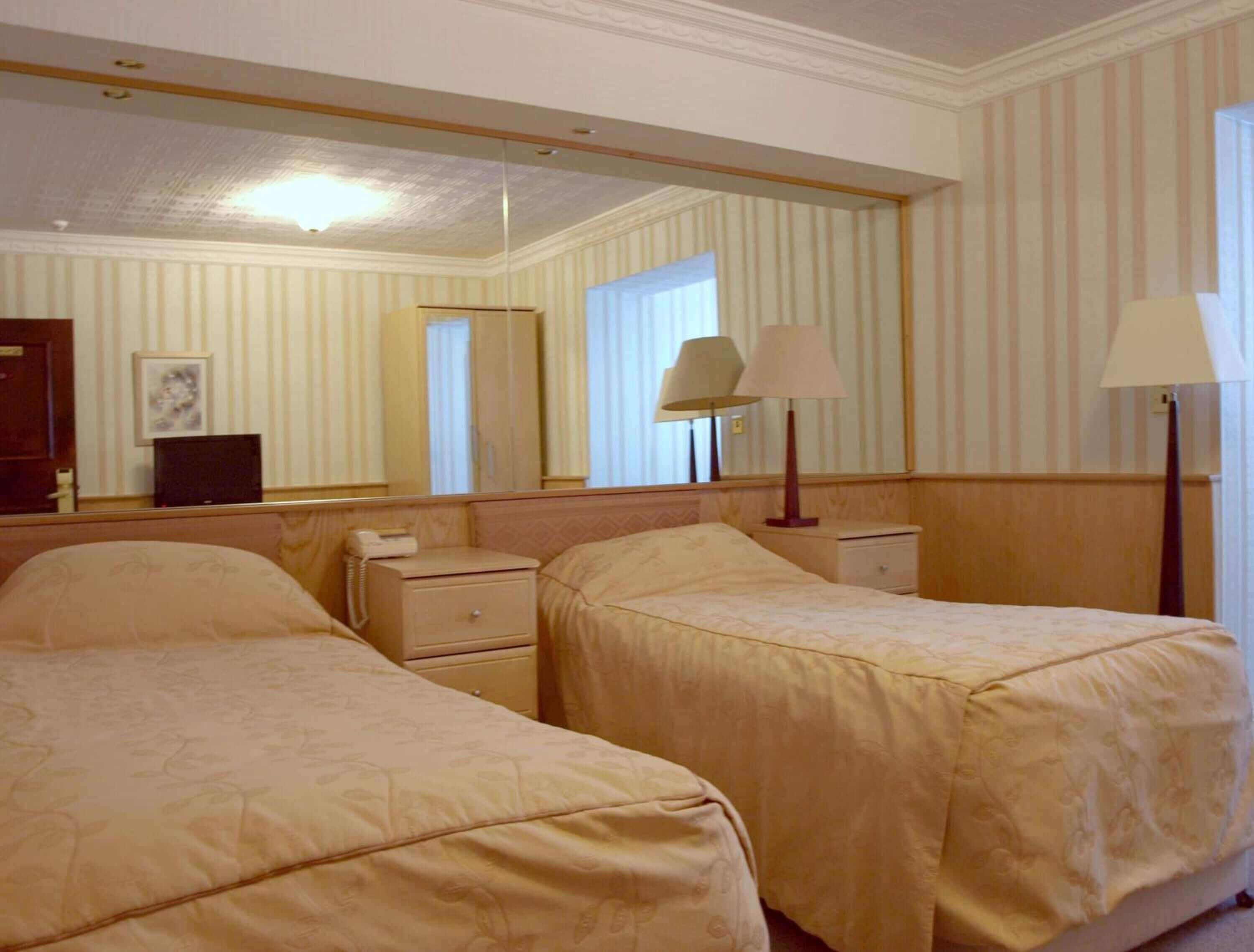 A hotel room with two beds, a bedside table with a lamp, a mirror, and a window with curtains.