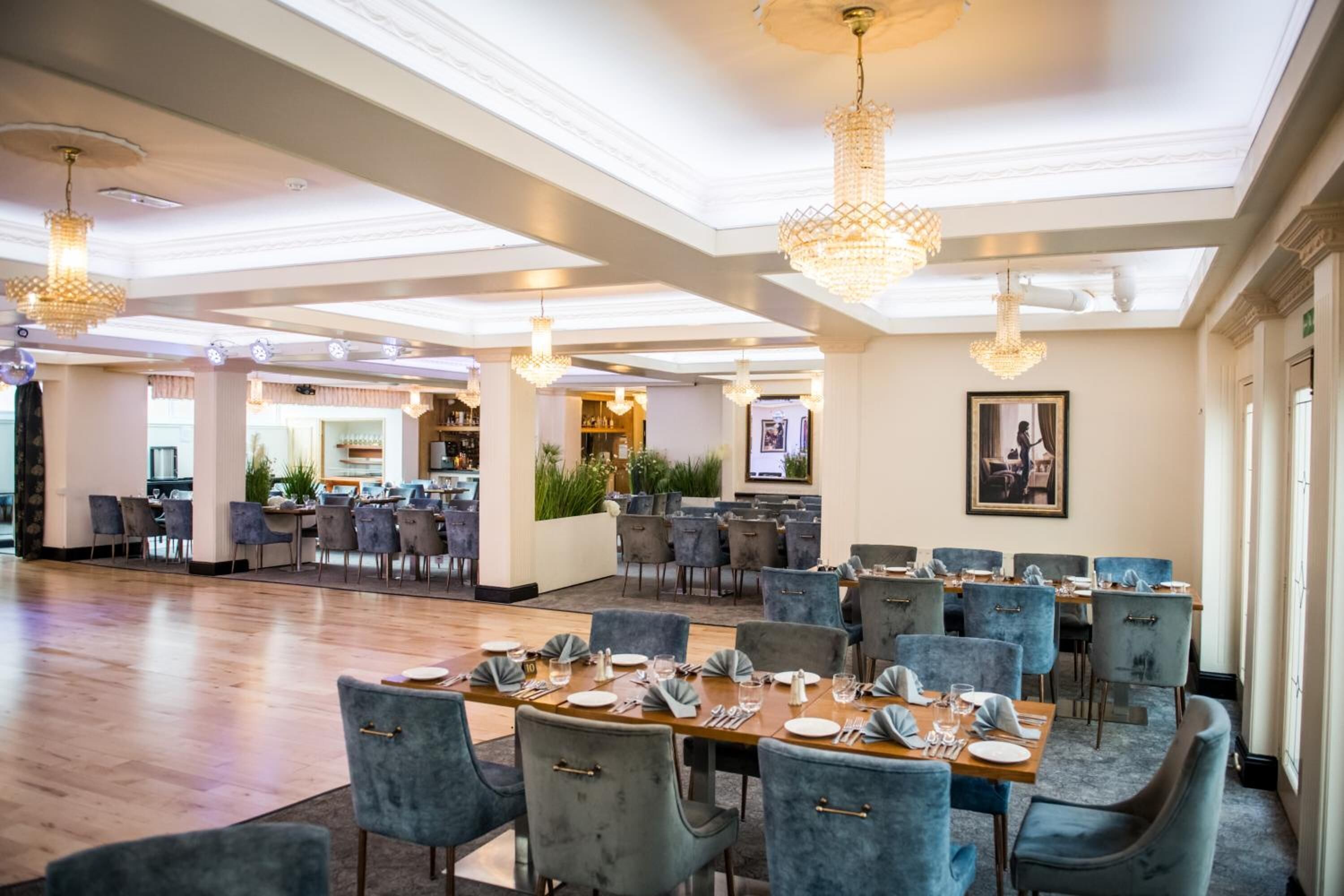 A spacious dining area with multiple tables and chairs, chandeliers, and a view of the kitchen in the background.