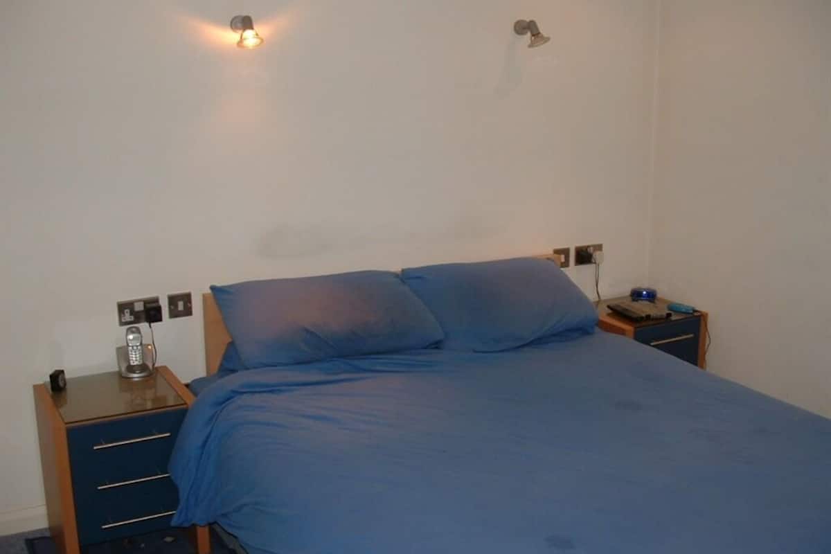 A single bed with a blue mattress and pillows, a bedside table with a lamp, and a power outlet on the wall.