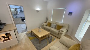 Un salón moderno con un sofá beige, una mesa de centro de madera y una cocina visible al fondo.
