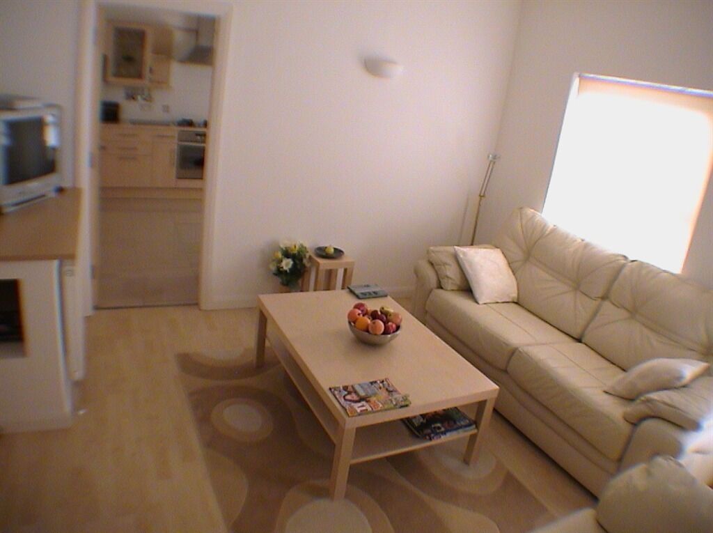 Un salon avec un canapé beige, une table basse sur laquelle se trouve un saladier rempli de fruits et un magazine. On aperçoit une cuisine au fond.