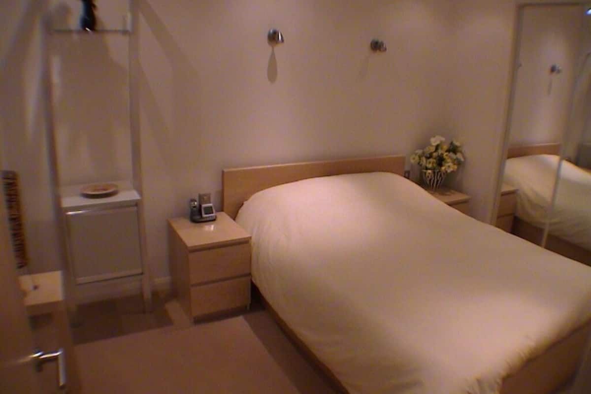 A hotel room with a single bed, a bedside table with a lamp, a small bedside table with a vase of flowers, and a mirror on the wall.