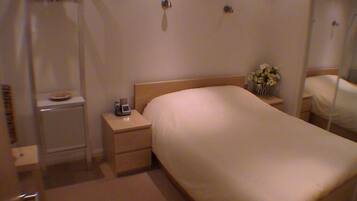 Une chambre d’hôtel avec un lit simple, une table de chevet avec une lampe, une petite table de chevet avec un vase de fleurs et un miroir au mur.