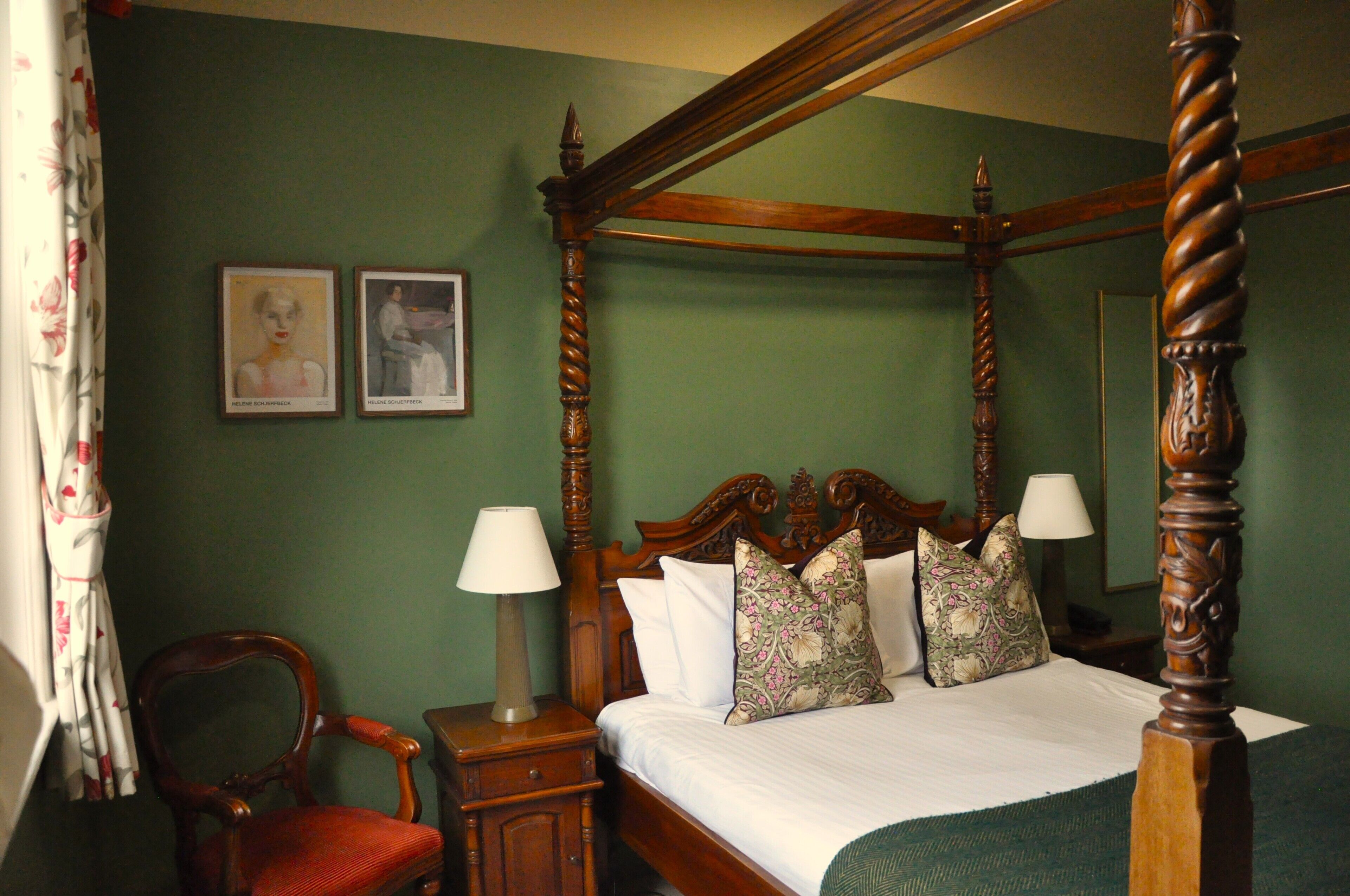 A four-poster bed with a wooden frame, a green wall, two framed artworks, a chair, and a nightstand with a lamp.