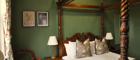 A four-poster bed with a wooden frame, a green wall, two framed artworks, a chair, and a nightstand with a lamp.