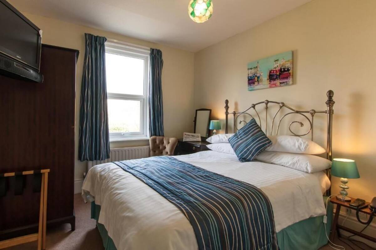 A bedroom with a bed, a nightstand, a lamp, a dresser, a window with curtains, and a painting on the wall.