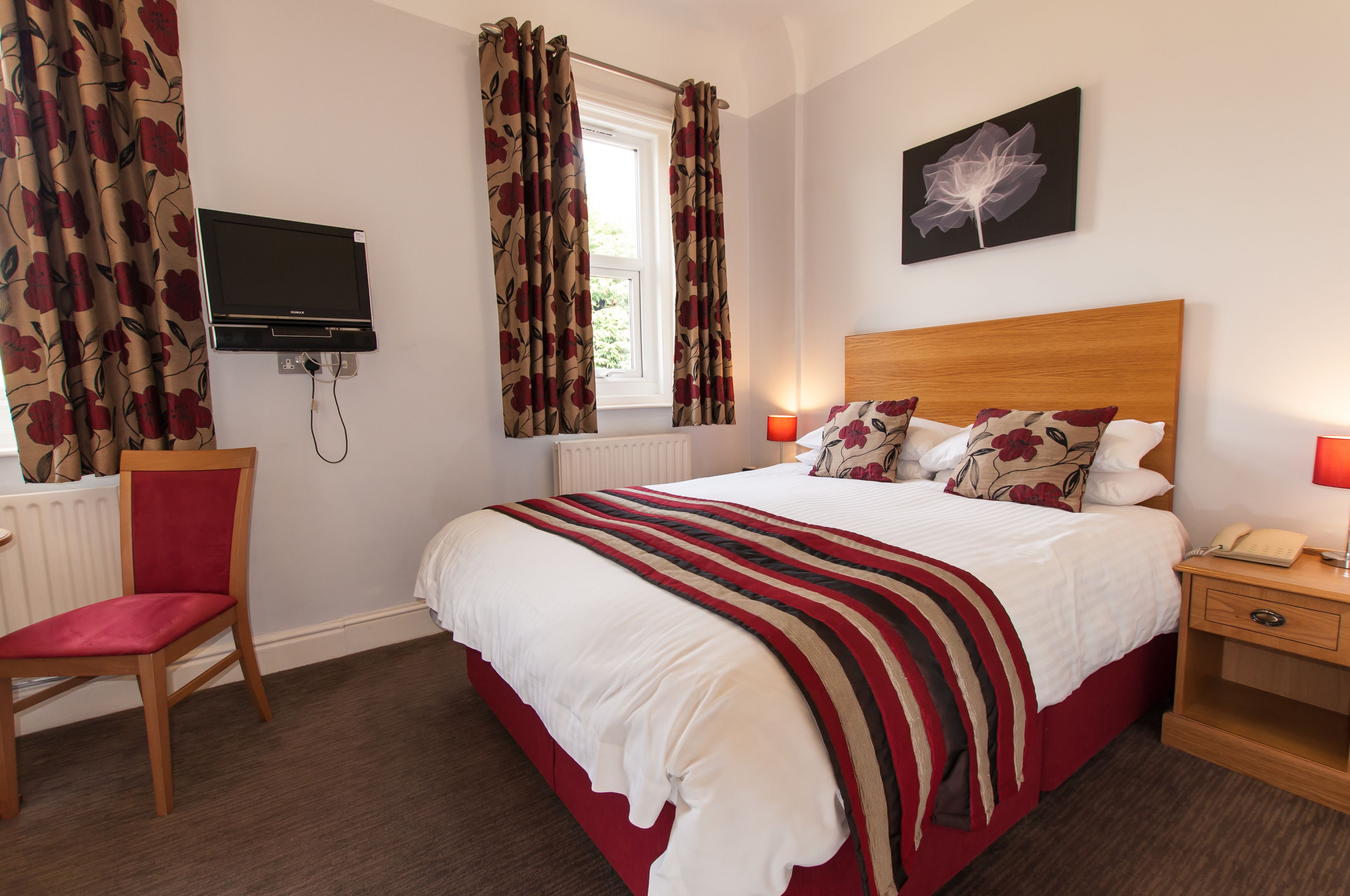 A hotel room with a bed, a chair, a TV, a window with curtains, a bedside table with a lamp, and a wall-mounted artwork.