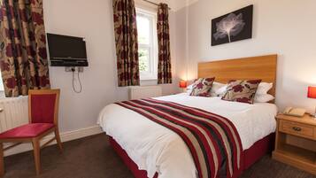 A hotel room with a bed, a chair, a TV, a window with curtains, a bedside table with a lamp, and a wall-mounted artwork.